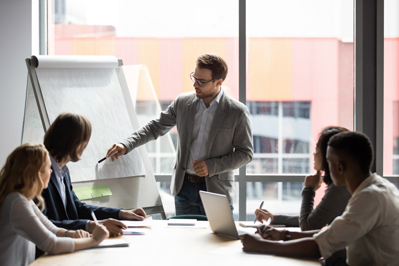 Business professional presenting ideas to a team during a meeting in a modern office conference room with a whiteboard and laptops