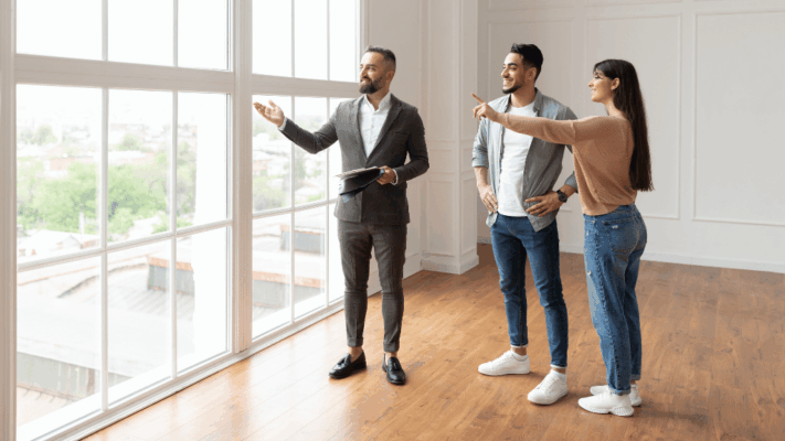 A real estate agent showing a modern luxury apartment to a young couple during a property tour, featuring bright natural light, large floor-to-ceiling windows, hardwood flooring, and upscale interiors. The scene represents advanced capital allocation for passive investors in Albany NY, multifamily real estate investing, property acquisition strategy, investor decision-making, high-end residential real estate opportunities, wealth-building through passive investing, real estate portfolio growth, and professional property consultation.
