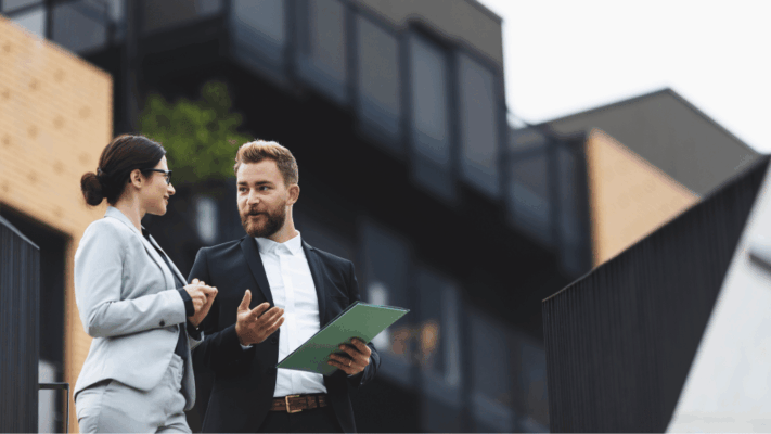Two business professionals discussing commercial real estate and investment opportunities outside a modern apartment or multifamily property development. The image represents real estate syndication, property underwriting, investment analysis, multifamily housing strategies, commercial property evaluation, professional consulting, real estate partnerships, and advanced deal assessment for property investors.
