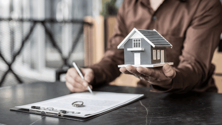 A professional real estate finance scene featuring a person reviewing and signing documents while holding a miniature house model, symbolizing analyzing debt structures in Albany NY syndication opportunities. The image represents commercial real estate financing, passive investment analysis, mortgage and loan structuring, real estate syndication evaluation, investment risk assessment, multifamily property funding strategies, capital management, and financial due diligence for institutional-grade real estate deals.