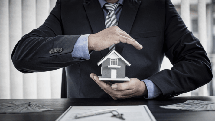 Business professional shielding a miniature house model with both hands over financial documents and cash, symbolizing building a risk-adjusted real estate strategy, asset protection, investment risk management, property portfolio security, real estate financial planning, wealth preservation, commercial property investing, insurance and liability protection, strategic investment analysis, and long-term real estate stability.