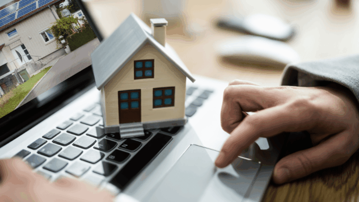 Model house placed on a laptop keyboard while a person researches real estate investment opportunities online