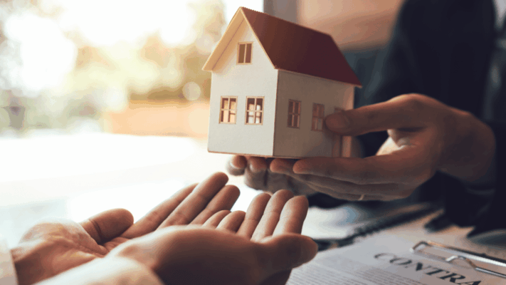 A warm, professional real estate transaction scene showing a house model being handed over between two people, symbolizing inflation hedging through passive real estate in Albany NY. The image highlights passive income investing, wealth preservation, real estate syndication, long-term asset protection, multifamily investment strategies, financial security during inflation, property ownership transfer, and stable real estate-backed investments.