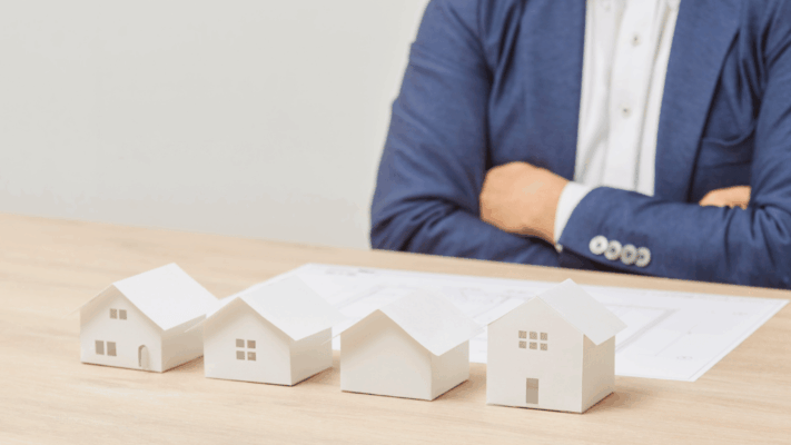 Business professional sitting behind a row of miniature house models on a desk, symbolizing leveraging market cycles in real estate investing, property portfolio management, housing market trends, strategic real estate planning, investment timing, residential property analysis, market diversification, real estate forecasting, long-term investment strategies, and wealth building through property investments.
