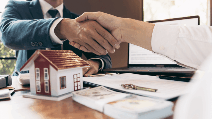 Real estate professionals shaking hands over a property investment agreement with a model home, financial documents, and cash on the desk 