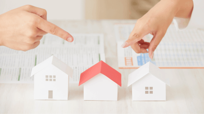 Hands pointing toward miniature model houses placed on financial documents, symbolizing multifamily real estate evaluation, property comparison, investment risk assessment, stress testing syndication deals, housing market analysis, real estate decision-making, property underwriting, financial planning, and strategic real estate investment discussions.
