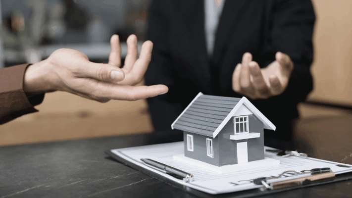 A real estate investment discussion scene featuring a miniature house model placed on top of documents while two people gesture during a financial conversation, symbolizing value creation metrics in Albany NY syndication deals. The image represents property performance analysis, passive real estate investing, multifamily syndication evaluation, investor strategy discussions, asset value growth, financial decision-making, commercial real estate metrics, and measuring returns in institutional-grade property investments.
