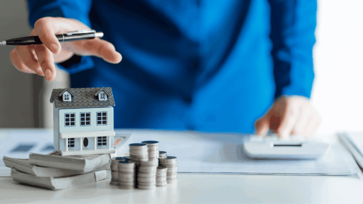 A professional real estate investment scene featuring a model home surrounded by stacks of cash and coins while an investor reviews financial information with a calculator. The image represents foundational real estate investing concepts, commercial real estate education, multifamily investing, passive income strategies, financial analysis, investment planning, property valuation, underwriting, portfolio building, and the essential real estate investment terms every Albany NY investor should understand before evaluating opportunities.