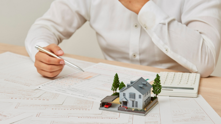A person reviews property-related documents with a pen in hand while a detailed model home and calculator sit on the desk, illustrating the ongoing analysis and oversight required to protect a real estate investment. The image conveys careful financial review, strategic planning, and performance monitoring, making it ideal for topics such as **how professional asset management protects real estate investments**, multifamily asset management, property performance analysis, investment oversight, financial reporting, risk management, operational efficiency, portfolio management, value-add strategies, and protecting long-term investor returns.
