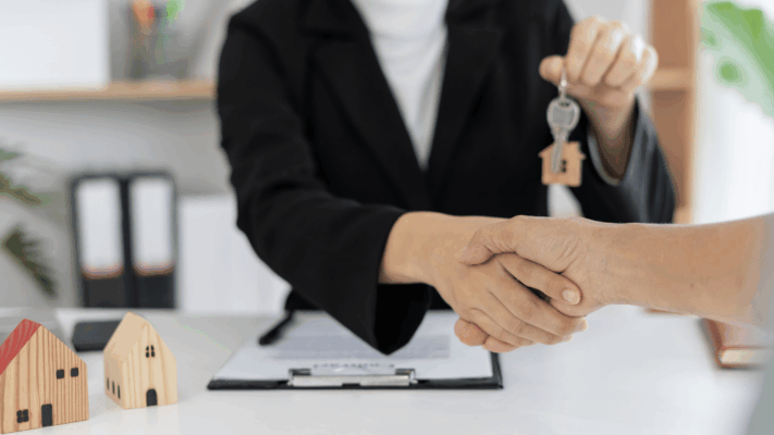 A professional real estate transaction scene featuring a handshake between an investor and an advisor while a house-shaped key is handed over, with miniature homes and contract documents in the background, symbolizing scaling from 10 to 100 units in the Albany NY market. The image represents multifamily portfolio growth, passive real estate investing, investment partnerships, commercial property expansion, real estate syndication, portfolio scaling strategies, long-term wealth building, and successful acquisition of larger real estate assets in Albany NY.