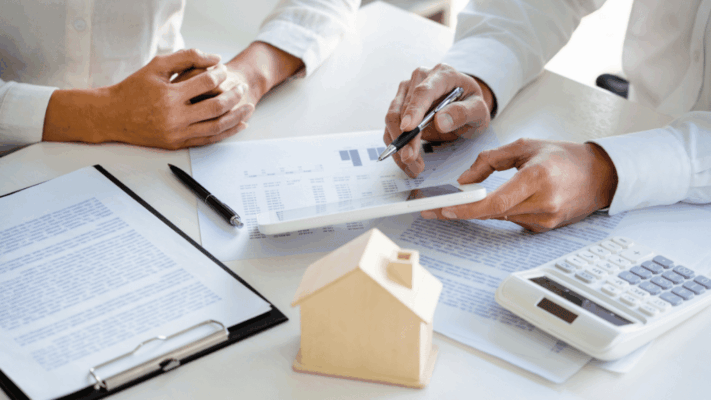 A professional real estate investment planning meeting featuring advisors reviewing financial reports on a tablet alongside investment documents, a calculator, and a miniature house model, symbolizing timing entry points in Albany NY syndication opportunities. The image represents passive real estate investing, market timing strategies, real estate syndication analysis, multifamily investment planning, financial due diligence, investment decision-making, portfolio optimization, market cycle evaluation, and identifying optimal opportunities for long-term commercial real estate growth.