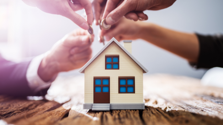 A miniature house sits in the foreground while several hands exchange coins and keys above it, symbolizing the collaborative management of a real estate investment. The image represents investors, sponsors, and property managers working together to maximize the value and performance of a shared asset. It is ideal for topics such as **understanding asset management in real estate syndications**, multifamily syndications, investor relations, property operations, portfolio management, cash flow optimization, value-add strategies, real estate partnerships, investment performance, and long-term asset management.

