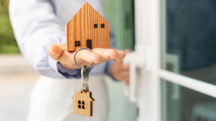 A person presents a wooden model home while holding a house-shaped keychain, symbolizing ownership, responsibility, and the ongoing management of a real estate investment. The image conveys the financial and operational commitment required to maintain a property's value, making it well suited for topics such as **understanding multifamily property operating expenses**, operating cost management, property maintenance, asset management, budgeting, multifamily ownership, expense tracking, cash flow management, investor returns, and maximizing long-term property performance. 