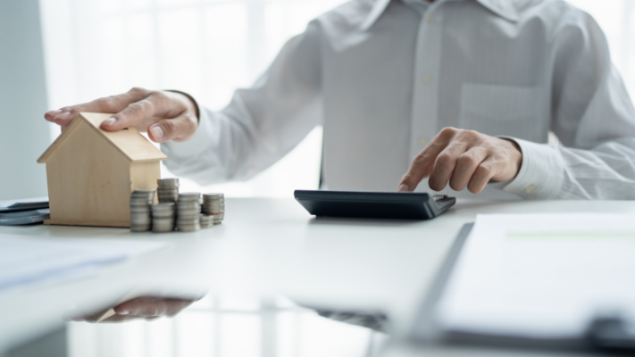 A person uses a calculator while resting a hand on a wooden house model beside neatly stacked coins, emphasizing careful financial planning and property investment analysis. The image is well suited for topics such as **understanding off-market multifamily opportunities**, real estate investment analysis, multifamily acquisitions, property valuation, due diligence, deal sourcing, investment strategy, financial planning, cash flow evaluation, and identifying hidden real estate opportunities before they reach the public market.