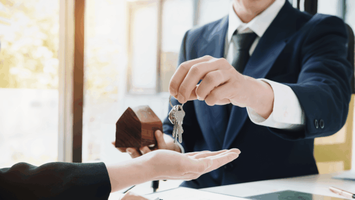 A professional real estate investment scene featuring an investor handing over a set of property keys while holding a model home, symbolizing ownership, investment structure, and capital allocation. The image represents preferred equity versus common equity, multifamily real estate syndications, passive investing, investor distributions, ownership interests, investment priorities, risk and return, commercial real estate partnerships, capital stack structure, and understanding equity positions in Albany NY syndications.