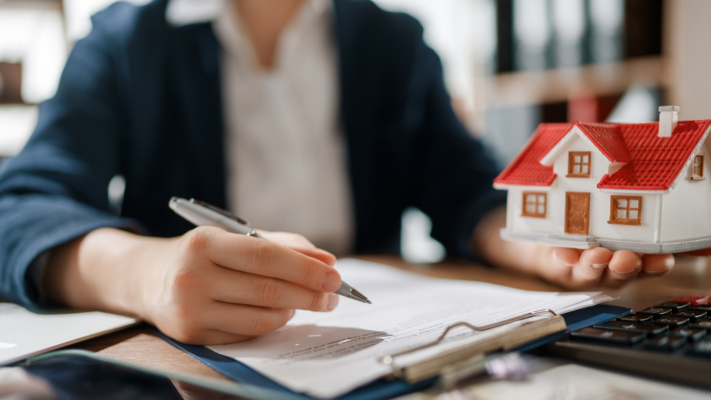 A real estate professional reviews documents while holding a model home, illustrating the ongoing oversight and strategic management that take place after an investment is made. The image conveys patience, performance monitoring, and long-term planning rather than a property sale, making it ideal for topics such as **what passive investors should expect during a hold period**, multifamily syndications, asset management, investor updates, property performance, business plan execution, cash flow distributions, portfolio monitoring, long-term wealth building, and maximizing investment returns over the life of a real estate investment.
