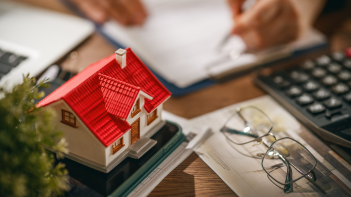 A model home with a bright red roof sits on a desk surrounded by financial documents, a calculator, and reading glasses while someone reviews paperwork in the background. The image represents the financial and operational analysis required to keep multifamily properties performing at their best. It is ideal for topics such as why occupancy rates matter in multifamily investing, occupancy and vacancy analysis, rental income stability, cash flow management, property performance, multifamily asset management, financial planning, investor returns, leasing strategies, tenant retention, and maximizing long-term real estate investment success.